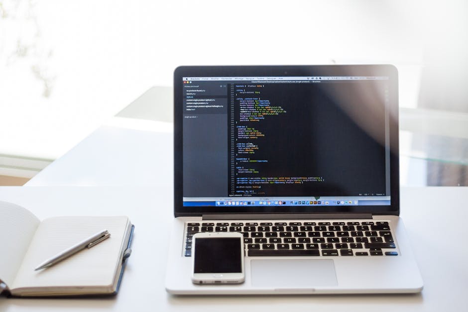 A close-up view of a modern workspace featuring a silver MacBook laptop displaying lines of code on its screen, positioned on a white desk. To the left of the laptop, an open blank notebook with a sil